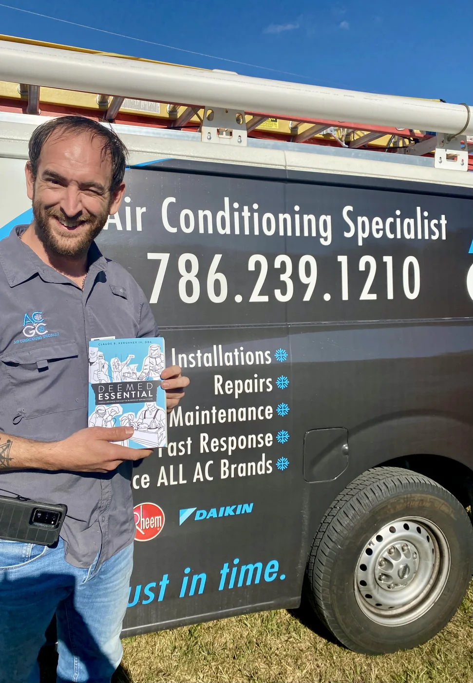 An HVAC professional holding a copy of Deemed Essential beside a service vehicle